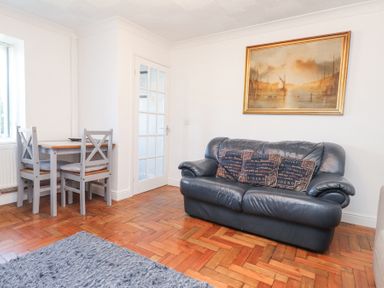A living room with a black leather sofa, three cushions, a small wooden dining table with four gray chairs, a large framed painting on the wall and a gray rug on the wooden floor at Tanyfron Holiday Studio in Bryngwran near Gwalchmai