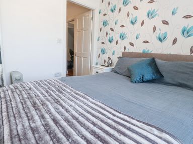A bedroom with a bed with blue bedding and a white bedside table near a door with floral wallpaper at Tanyfron Holiday Studio Bryngwran near Gwalchmai