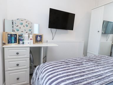A bedroom with a dresser with toiletries and framed pictures a wall-mounted TV a radiator and a wardrobe at Tanyfron Holiday Studio in Bryngwran near Gwalchmai