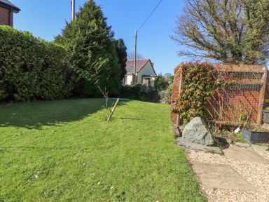 A garden with grass, trees, a wooden fence, a rock, and a house in the background at Tanyfron Holiday Studio in Bryngwran near Gwalchmai