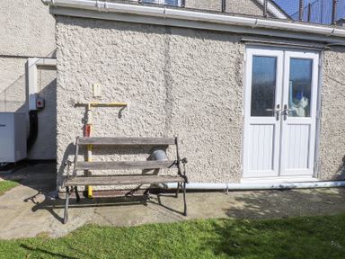 A wooden bench on a concrete patio in front of a textured wall with a double glass door and visible pipes at Tanyfron Holiday Studio in Bryngwran near Gwalchmai