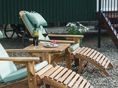 Wooden lounge chairs with light green cushions and a small table holding a bottle of wine wine glasses and a plate with food on gravel at Onnen Oakford near Aberaeron
