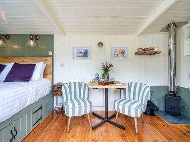 A small room with two striped chairs around a wooden table next to a bed and a wood stove at Onnen in Oakford near Aberaeron