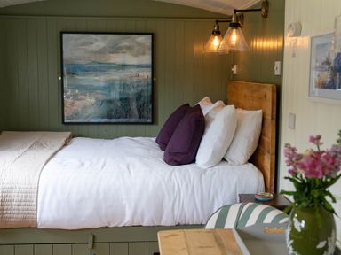 A bedroom with a bed with white sheets and purple pillows a wooden headboard a painting on the green paneled wall and a table with flowers at Onnen in Oakford near Aberaeron