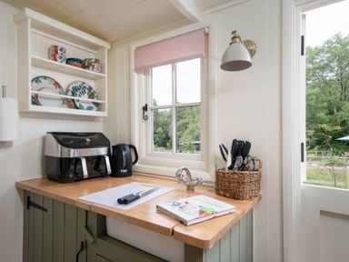 A kitchen countertop with a cutting board and knives a black kettle a wicker basket with utensils an air fryer and floral plates on a wall shelf at Onnen Oakford near Aberaeron