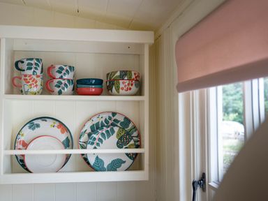 A kitchen shelf with cups bowls and plates with leaf patterns near a window with a pink blind at Onnen Oakford near Aberaeron