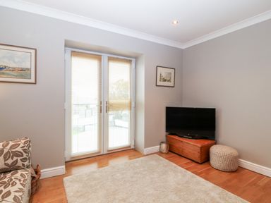 A living room with a television on a wooden chest and French doors with blinds at 10 Pen Llanw Tides Reach in Rhosneigr
