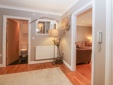 A hallway with wooden floor a carpet floor lamp a mirror and open doors showing a bathroom and a living room at 10 Pen Llanw Tides Reach in Rhosneigr