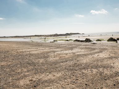 A sandy and rocky beach with people near water and distant land at 10 Pen Llanw Tides Reach in Rhosneigr