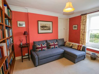 A living room with a gray sectional sofa with Union Jack pillows a wooden bookshelf and a small side table with a lamp at The White House in Cartmel