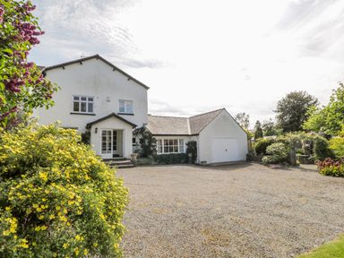 A white two-story house with attached garage and gravel driveway surrounded by bushes and trees at The White House in Cartmel