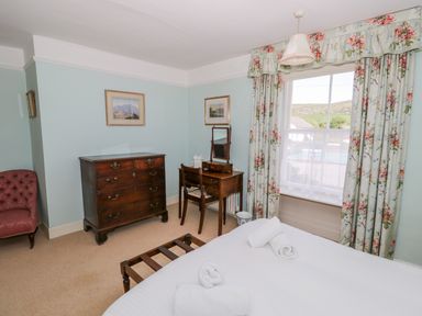 A bedroom with a bed with rolled towels wooden chest of drawers wooden dressing table with chair floral curtains and framed pictures at The White House in Cartmel