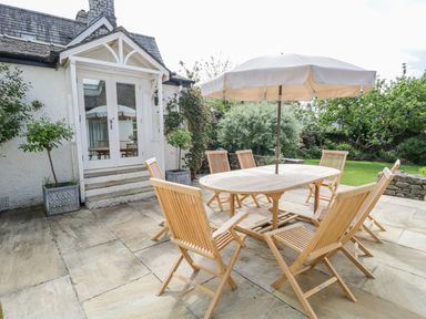An outdoor patio with wooden table and chairs and an umbrella near a white house with garden at The White House in Cartmel
