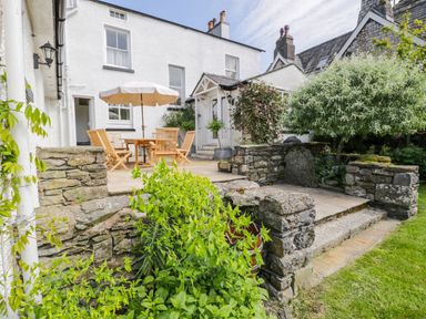 An outdoor patio with stone walls wooden table and chairs and a beige umbrella in a garden at The White House in Cartmel