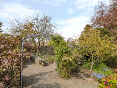 A garden pathway with plants climbing a metal arch and various shrubs and flowers at The White House in Cartmel