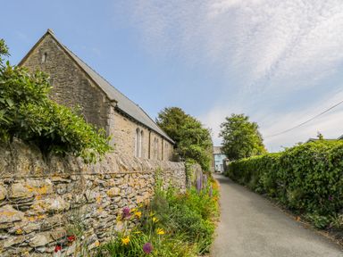 A narrow road lined with stone and ivy-covered walls with flowers and a stone building on one side at The White House in Cartmel