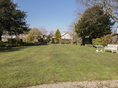 A garden with a lawn surrounded by trees shrubs and a stone wall with a white bench and table on the right at The White House in Cartmel