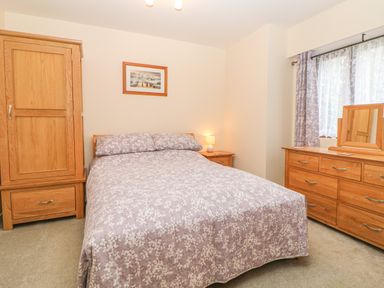 A bedroom with a double bed covered in floral bedding a wooden wardrobe bedside table with lamp and a wooden dresser with mirror at Hilly Field Barn in Yelverton