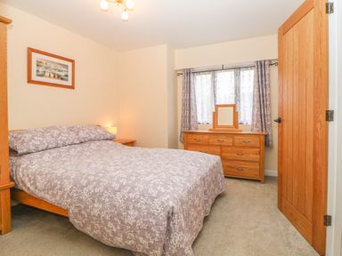 A bedroom with a double bed with floral bedding a wooden dresser with a mirror curtains on the window and a wooden door at Hilly Field Barn in Yelverton