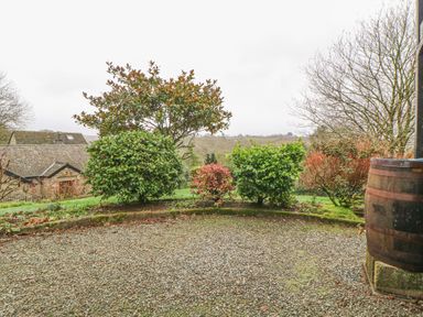 A garden area with bushes gravel ground and a wooden barrel near a stone building at Hilly Field Barn in Yelverton