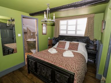 A bedroom with a carved wooden bed with patterned bedding and towels on it a window with shutters and patterned curtains and a door leading to a bathroom at The Old Paper Shop in Tewkesbury