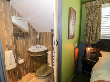 A small bathroom with a corner sink, toilet, towel rack, and wooden wall panels next to a bedroom with green walls, patterned curtains, and a bed at The Old Paper Shop in Tewkesbury