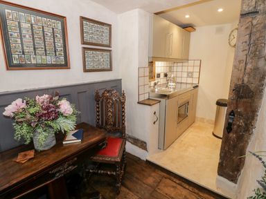 A small kitchen area with cabinets and sink next to a wooden table with flowers and a carved wooden chair at The Old Paper Shop in Tewkesbury