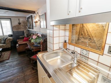 A kitchen sink with a wooden framed feature on the tiled wall next to a living area with chairs and a table with flowers at The Old Paper Shop in Tewkesbury
