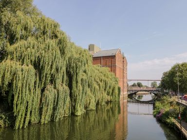 A river with large weeping willow trees a brick building and a bridge at The Old Paper Shop in Tewkesbury
