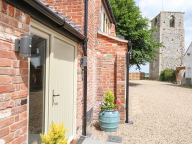 An exterior view of a brick building with a beige door and two potted plants near the entrance with a gravel driveway and a stone tower in the background at Cobblers Barn in Repps With Bastwick near Potter Heigham