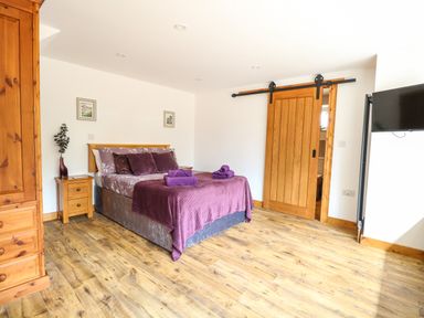 A bedroom with a double bed covered with purple bedding two wooden nightstands a wooden wardrobe a sliding wooden door and a wall-mounted television at Cobblers Barn in Repps With Bastwick near Potter Heigham