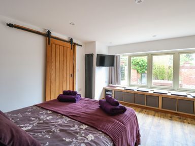 A bedroom with a bed covered in floral and purple bedding and folded towels a sliding wooden door a wall-mounted TV and a window bench with cushions at Cobblers Barn in Repps With Bastwick near Potter Heigham