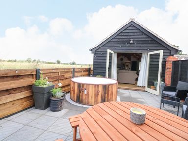 An outdoor patio area with a wooden table, hot tub, potted plants, and chairs outside a black wooden building with open doors at Cobblers Barn in Repps With Bastwick near Potter Heigham