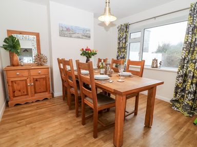 A dining room with wooden table and chairs a sideboard with a mirror and decorative items and a large window with patterned curtains at Cranberry in Bude