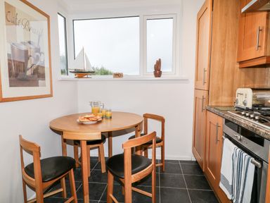 A kitchen dining area with a round wooden table and three chairs with black seats a plate of bread and glasses of juice on the table window with sailboat model and wooden decor on the windowsill wooden kitchen cabinets and a stove with a striped towel at Cranberry in Bude