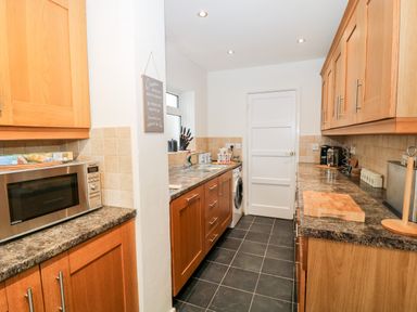 A kitchen with wooden cabinets black tiled floor a microwave a washing machine and utensils on the counter at Cranberry in Bude