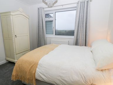 A bedroom with a large bed with white bedding and a beige throw next to a window with grey curtains and a cream wardrobe in Cranberry Bude