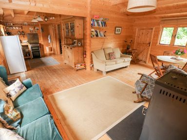 A wooden living room with green and beige sofas a wooden cabinet a round table with chairs and kitchen area at The Beach Shack in Llansteffan