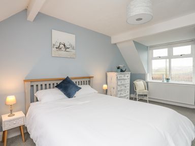 A bedroom with a bed a nightstand two lamps a chest of drawers a chair and a window at The Granary - Rhoscolyn