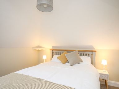 A bedroom with a bed with white bedding and two cushions two bedside tables with lamps and a wall shelf at The Granary - Rhoscolyn in Rhoscolyn