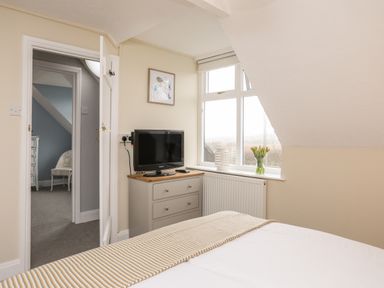 A bedroom with a bed a dresser with a television and a window with flowers on the sill at The Granary - Rhoscolyn in Rhoscolyn