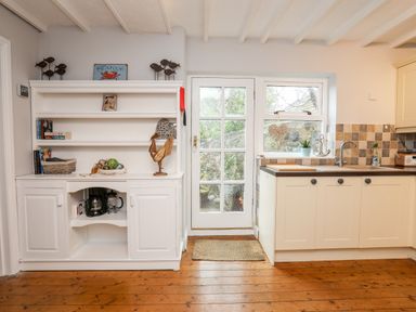 A kitchen area with white cabinets a wooden floor a white hutch with books and decorations and a glass door leading outside at The Granary - Rhoscolyn in Rhoscolyn