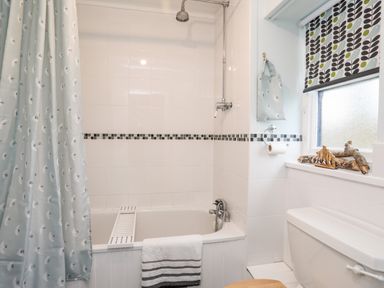 A bathroom with a bathtub shower combination a shower curtain a towel a window with patterned blinds and a toilet at The Granary - Rhoscolyn