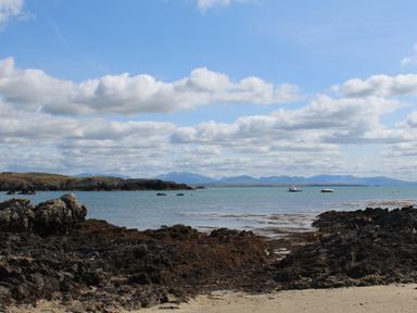 A rocky shoreline with seaweed and two boats on water with hills in the distance at The Granary - Rhoscolyn in Rhoscolyn