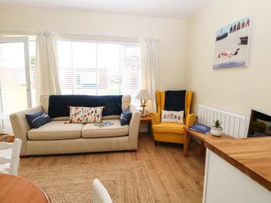 A living room with a beige sofa and yellow armchair near a window with white curtains at The Cranny in Mundesley