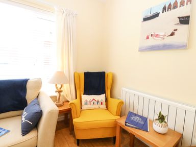 A corner with a yellow armchair a beige sofa side table with a lamp another side table with a plant and book and a painting on the wall at The Cranny in Mundesley