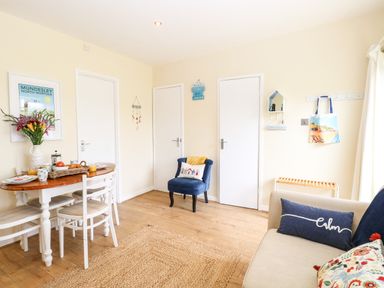 A dining area with a wooden table, white chairs, a blue armchair, a beige sofa with cushions and three doors in a room at The Cranny in Mundesley