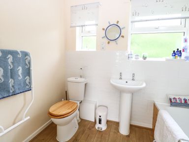 A bathroom with a toilet with a wooden seat a pedestal sink and a bathtub with towels and nautical decor at The Cranny in Mundesley