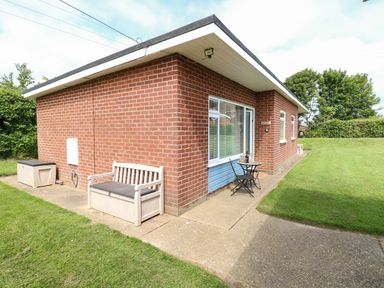 The outside corner of a brick house with a small bench and a table with two chairs on a concrete path surrounded by grass at The Cranny in Mundesley