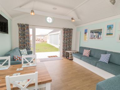 A living room with blue sofas and patterned cushions a wooden dining table white chairs and framed pictures on a light blue wall at Shore Thing 280 in Wilsthorpe